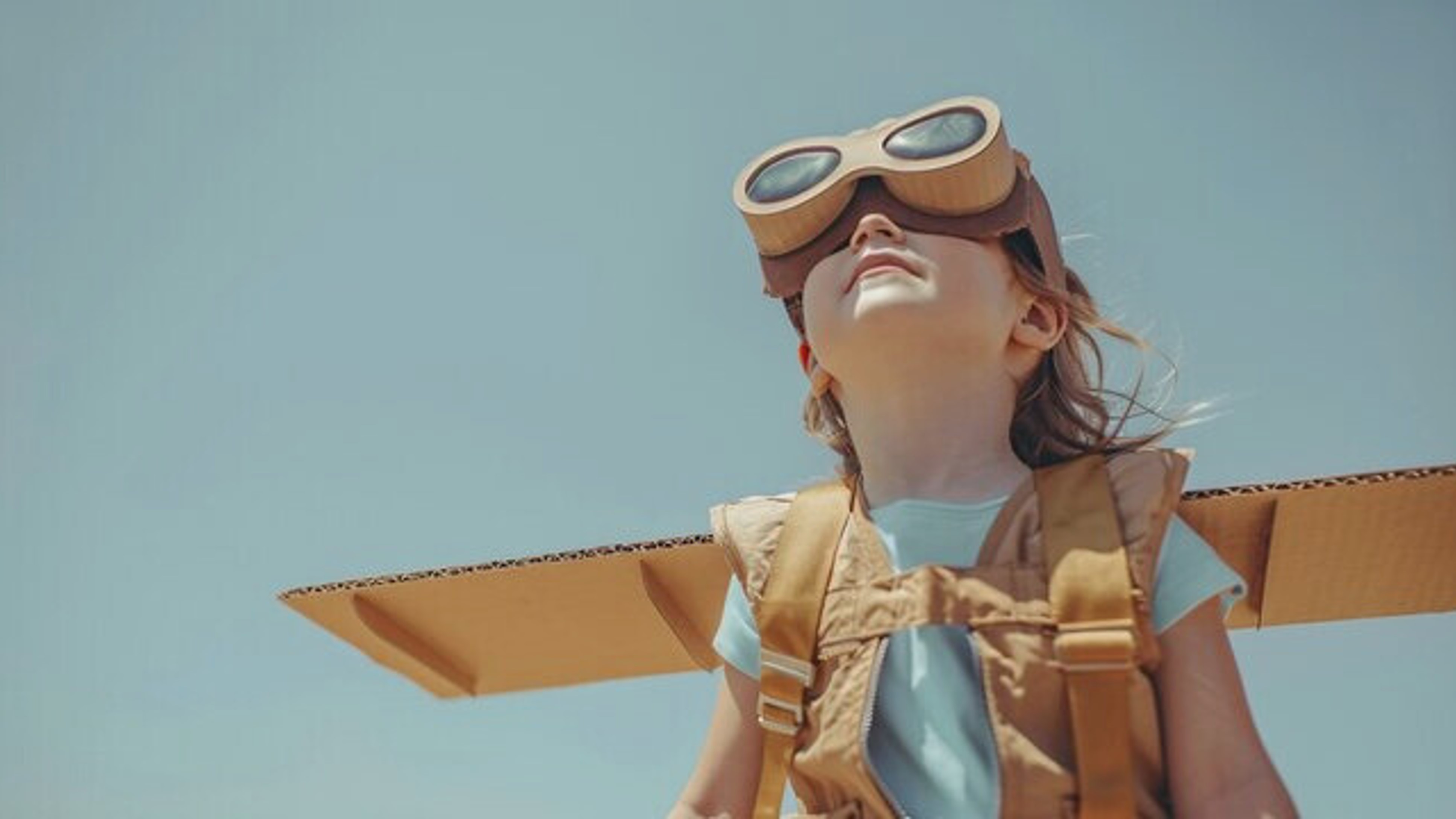 Un enfant portant des lunettes de pilote et un costume en carton, regardant vers le ciel avec un sourire, symbolisant l'imagination et la créativité qui font la force des enfants neuroatypiques