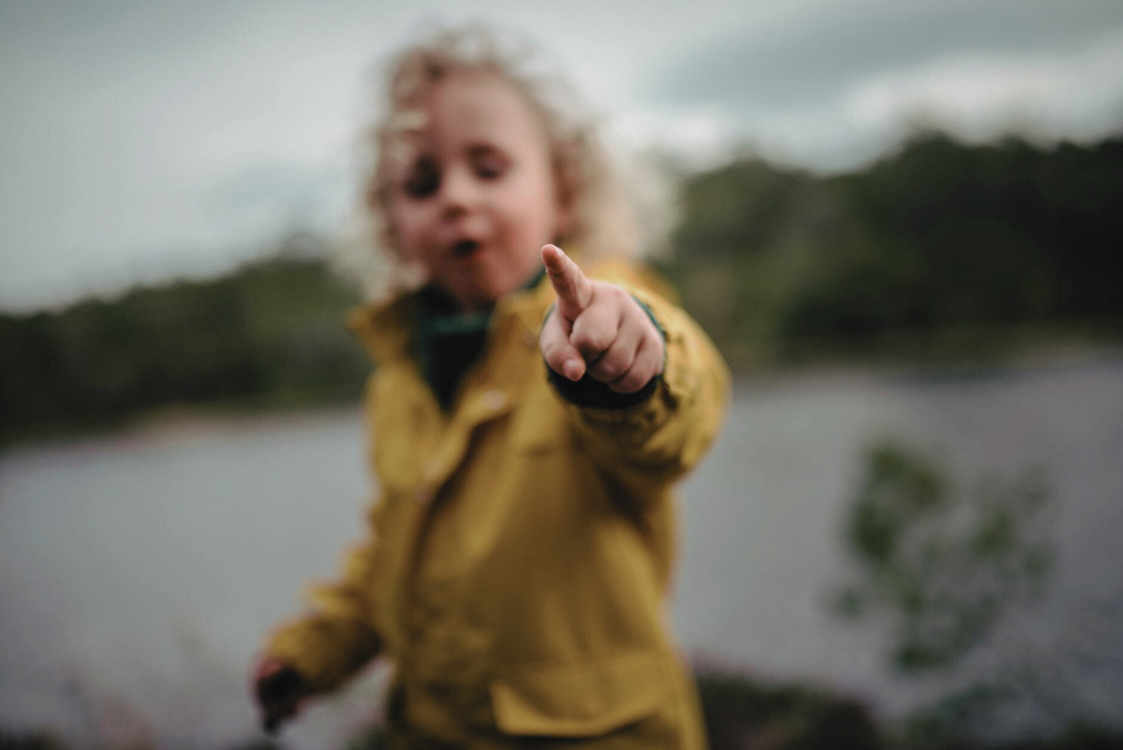 Un enfant aux cheveux bouclés portant un manteau jaune, pointant du doigt vers l'objectif, avec un paysage flou en arrière-plan.