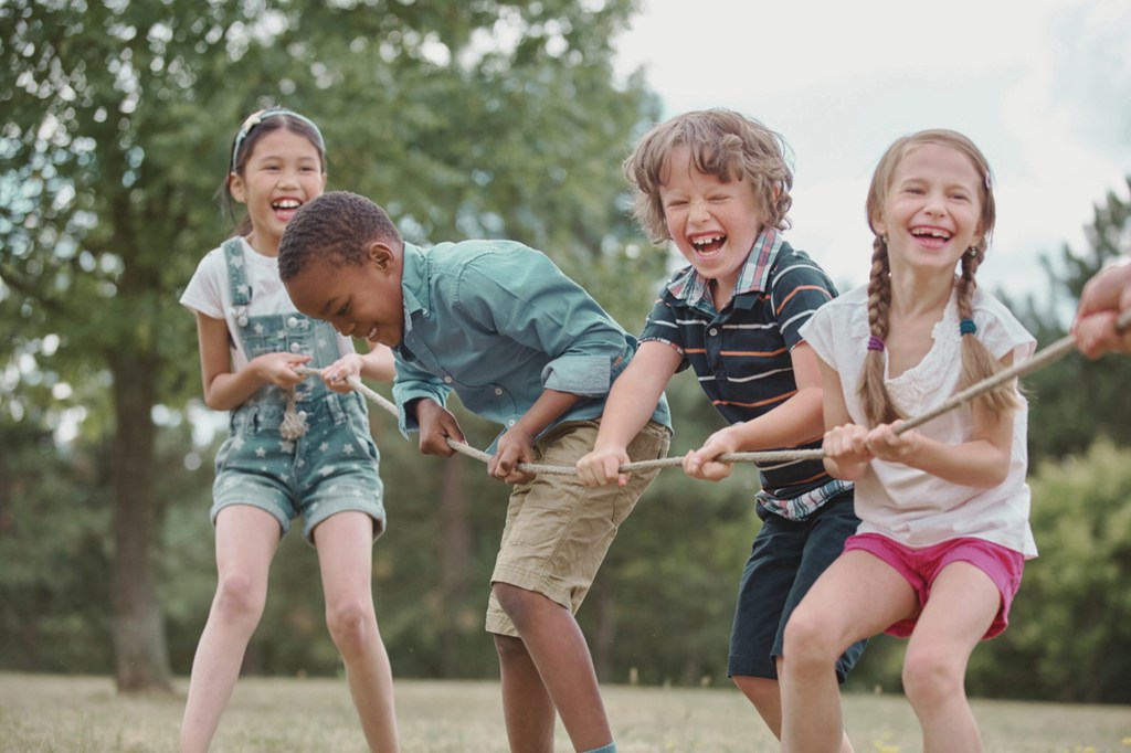 Quatre enfants s'amusent à tirer sur une corde dans un jeu en extérieur, riant et souriant, dans un environnement naturel.