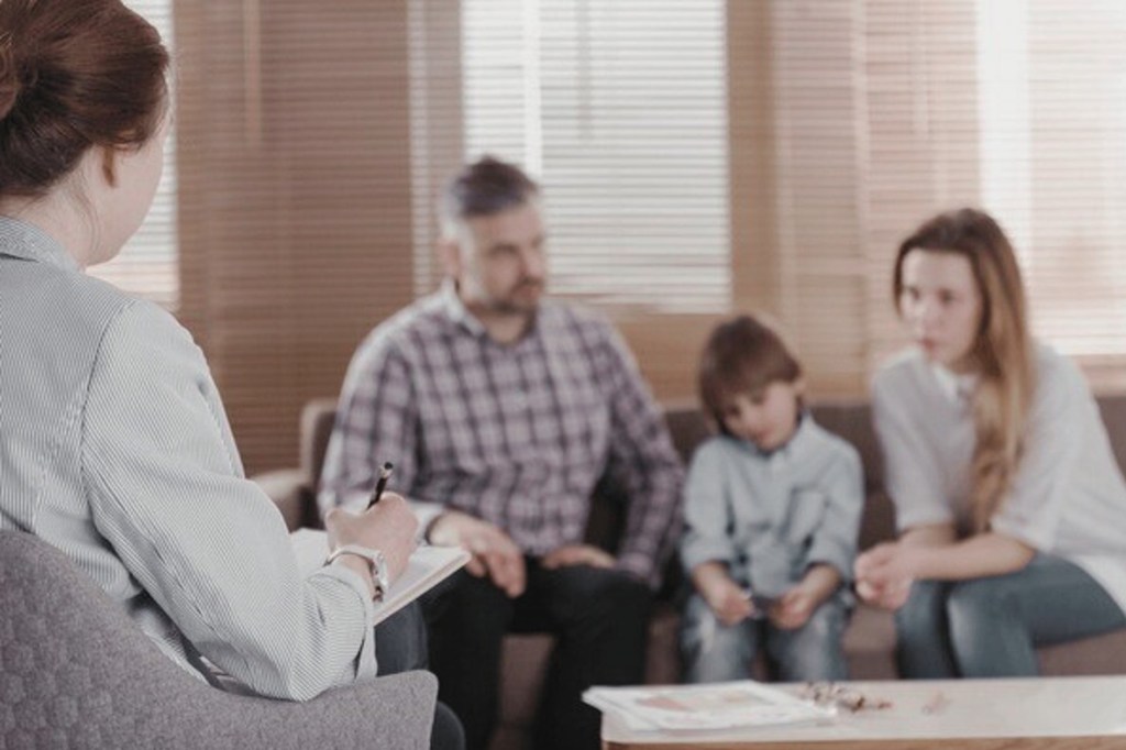 Un thérapeute observe et prend des notes lors d'une séance avec une famille, composée de deux adultes et un enfant, assis sur un canapé dans un environnement chaleureux.
